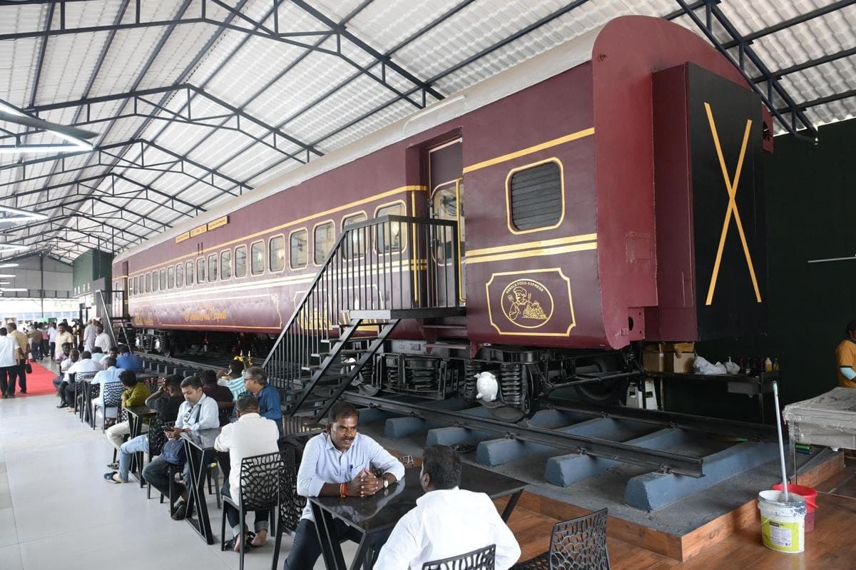 A maroon-and-gold heritage railway coach converted into a restaurant inside a shed, with steel stairs and diners at tables